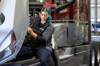 A Chemeketa Diesel student with blue hair is working on the engine of a large truck. They are wearing a black shirt with a logo on the chest and dark pants. The truck's hood is open, and the person is using a tool on the engine. The truck is red and silver with large tires. The scene appears to be in a garage or workshop with other vehicles and equipment in the background.